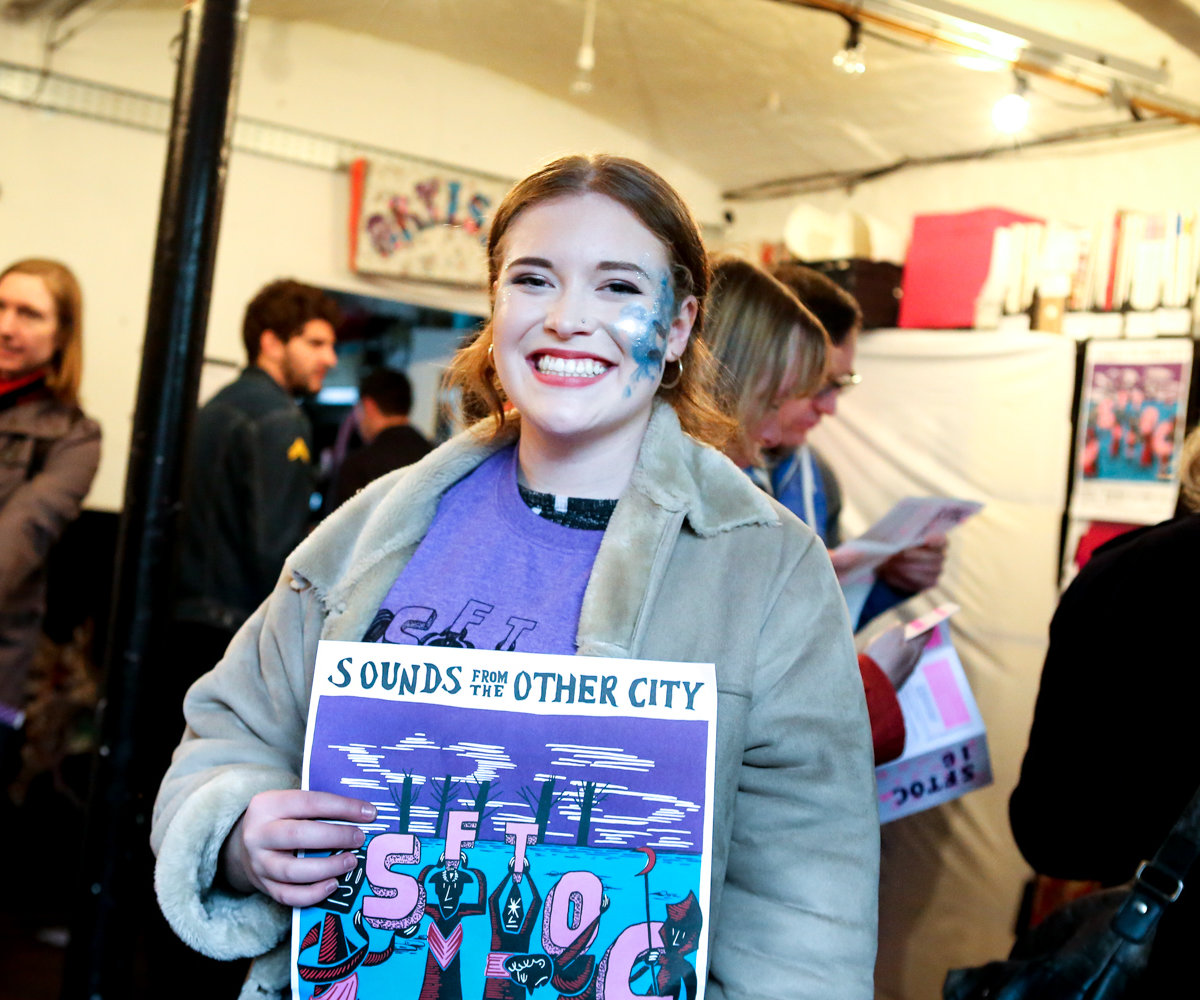 Photo of a female volunteer holding a SFTOC poster and smiling at the camera. She is white with brown hair tied back and blue glitter on her right cheek. She is wearing a beige jacket and purple tshirt.
