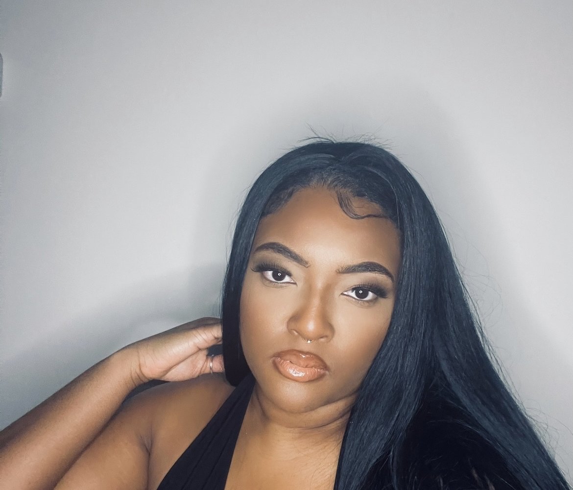 A woman stares directly into the camera, she wears all black and is against a grey wall